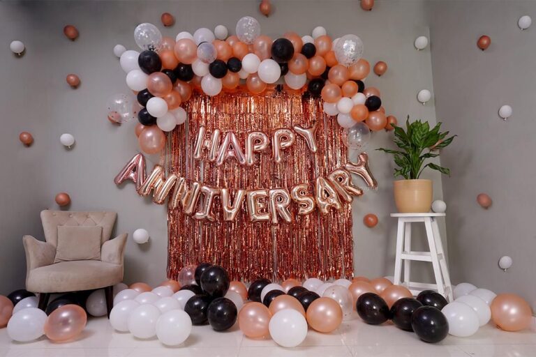 An elegant anniversary decoration setup against a grey wall. In the center, a rose gold foil curtain backdrop features large, matching "HAPPY ANNIVERSARY" foil balloons. This is framed by a beautiful balloon arch made of rose gold, black, white, and confetti-filled balloons. More balloons are scattered on the floor, and a plush beige armchair sits to the left, while a potted plant on a white stool is on the right.