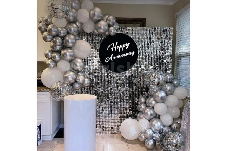 A modern and glamorous Sparkling Silver Anniversary Decoration Services NCR featuring a silver sequin shimmer wall backdrop. An elegant balloon garland with chrome silver, white, and confetti-filled balloons frames the scene. In the center of the shimmer wall is a circular black sign that reads "Happy Anniversary" in white script. A white cylindrical pedestal stands in the foreground.