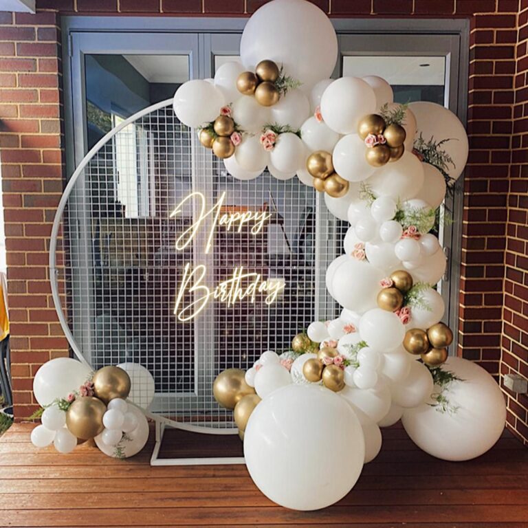 A luxurious, white and gold balloon garland forming an arch over a large circular mesh backdrop, featuring a neon 'Happy Birthday' sign in the center, set on a wooden deck in front of glass doors and a brick wall.