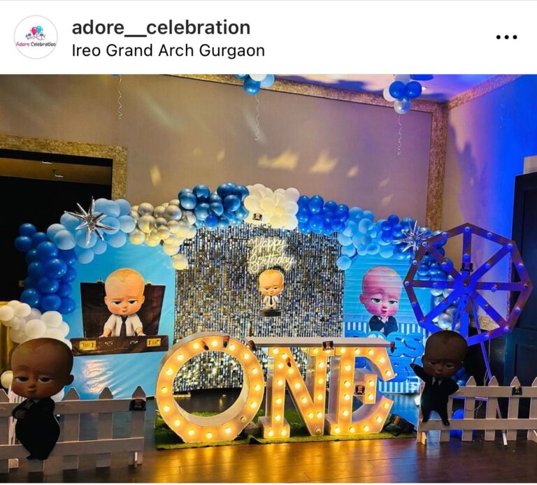 A vibrant Boss Baby themed birthday decoration featuring a blue and white balloon arch, a shimmery sequin backdrop, a neon "Happy Birthday" sign, and large, light-up marquee letters spelling "ONE." The setup includes Boss Baby character cutouts and a decorative white picket fence.