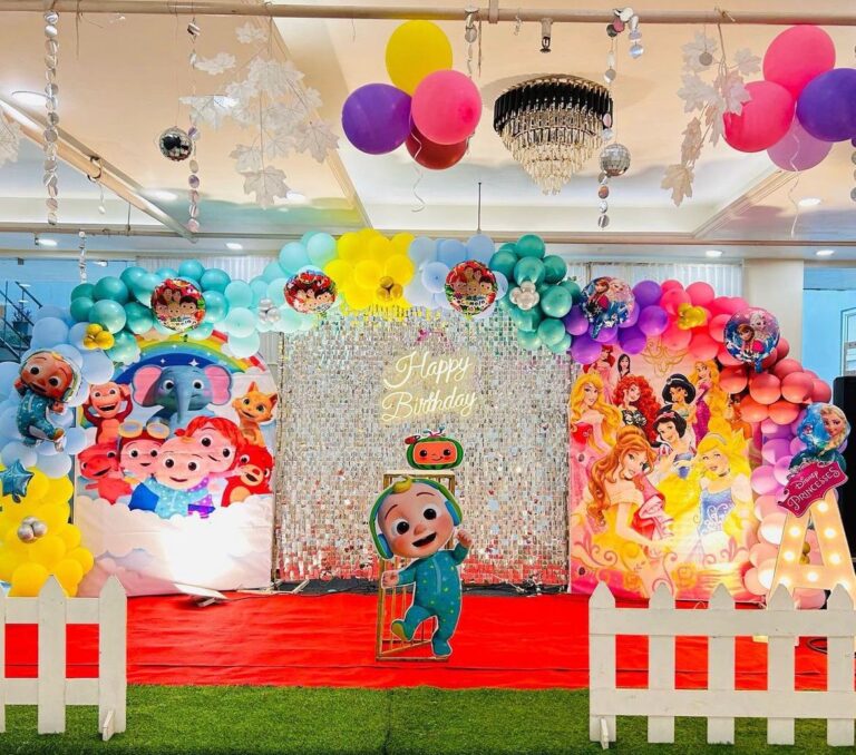 A vibrant Kids Cocomelon Theme Birthday Decor. On the left with JJ cutout, a silver sequin backdrop with "Happy Birthday" neon light, and a Disney Princess theme on the right with a large letter A. Colorful balloon arches frame both sides.