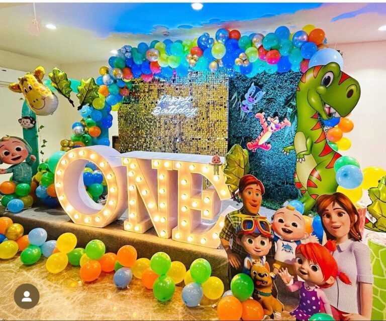 A vibrant indoor birthday party decoration with a colorful balloon arch, a large light-up "ONE" sign, and cutouts of Cocomelon characters and a dinosaur.