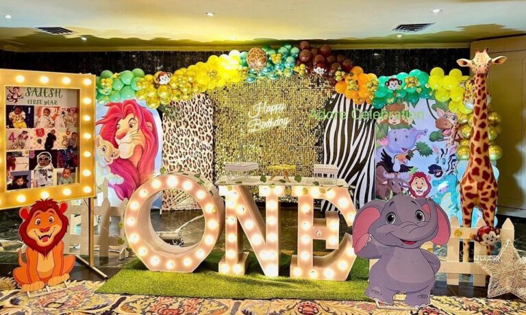 An elaborate jungle-themed first birthday party decoration. In the center is a large, illuminated marquee sign spelling "ONE". The backdrop features gold sequins, leopard and zebra prints, and is topped with a colorful balloon arch. Cardboard cutouts of characters like Simba and Mufasa from "The Lion King," an elephant, and a tall giraffe surround the scene. A large, lit-up photo frame on the left displays a collage of a baby's first year.