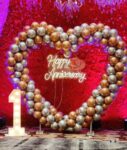 A large, heart-shaped balloon arch in metallic gold and silver with a neon "Happy Anniversary" sign inside and a glowing number "1" on the left.