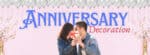 A banner image for "Anniversary Decoration" featuring a romantic couple sharing a moment, holding a heart-shaped gift between them. The background is a soft pink, adorned with silver balloons, golden confetti, and two champagne bottles at the bottom corners.