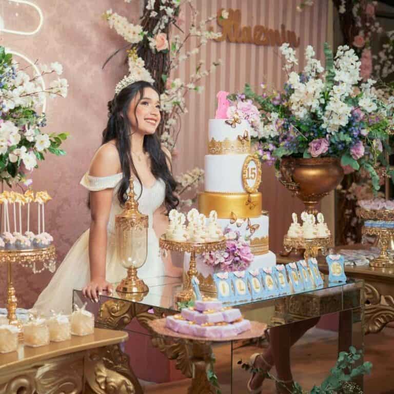 A smiling young woman wearing an off-white gown and tiara, standing beside a beautifully decorated dessert table at her 15th birthday (Quinceañera) celebration. The table features a tall multi-tiered cake with gold accents and butterflies, various pastries, and floral arrangements. The background is decorated with striped wallpaper and abundant flowers.