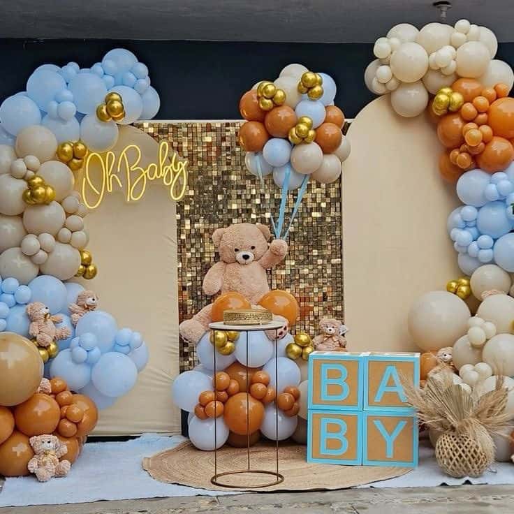 Elaborate welcome baby decoration featuring blue, white, and gold balloons forming arches, a sparkly gold backdrop with a neon "Oh Baby" sign, a large teddy bear holding balloons, and wooden blocks spelling "BABY".