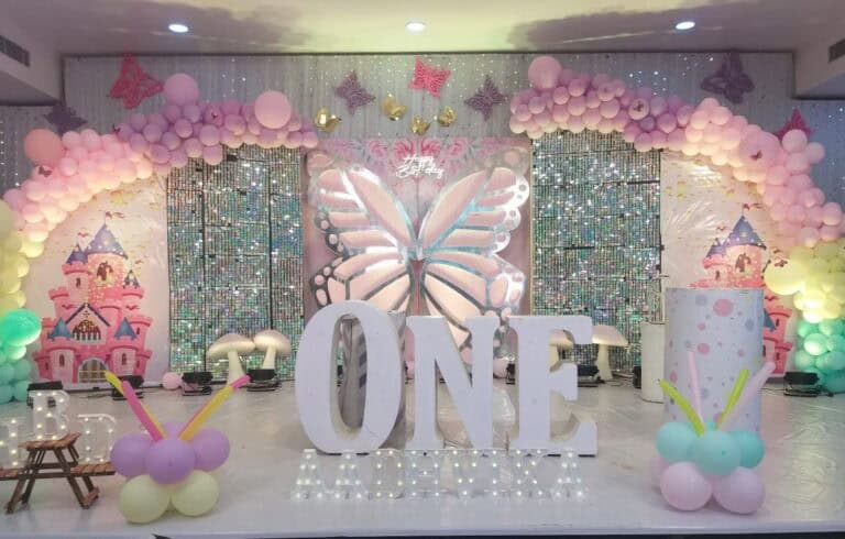 An elaborate first birthday party stage decoration with a butterfly and princess theme. The setup features a large, illuminated butterfly centerpiece, extensive pink and purple balloon arches, a shimmering sequin wall, and large light-up marquee letters spelling "ONE" in the foreground.