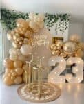 A stylish birthday party decoration setup featuring an organic balloon garland in chrome gold and shades of beige. In the center is a circular sign with a "Happy Birthday" neon light, flanked by three gold pedestal stands and large, illuminated marquee numbers showing "23" on a woven rug.