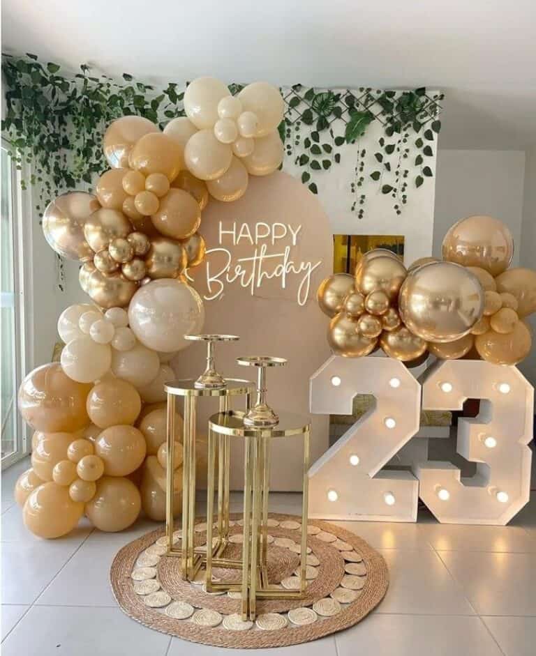 A stylish birthday party decoration setup featuring an organic balloon garland in chrome gold and shades of beige. In the center is a circular sign with a "Happy Birthday" neon light, flanked by three gold pedestal stands and large, illuminated marquee numbers showing "23" on a woven rug.