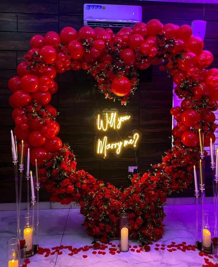 A large, vibrant red heart-shaped balloon and rose flower arrangement with a glowing neon sign in the center that reads "Will you Marry me?" surrounded by candles and rose petals on the floor.