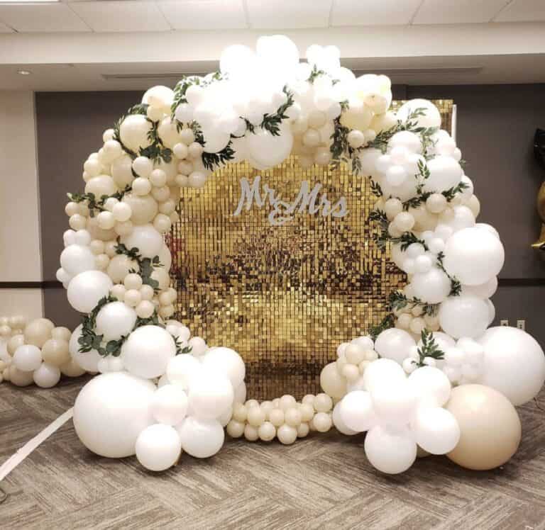 An elegant balloon ring decoration with white and cream organic balloons and greenery, framing a gold sequin shimmer wall with a white 'Mr & Mrs' sign at the center.