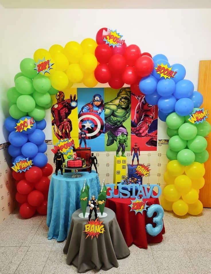 A complete superhero-themed birthday party decoration featuring a colourful balloon arch in red, blue, green, and yellow. The setup includes an Avengers backdrop, a cake table with action figures, and personalized name and age displays for a child's third birthday.