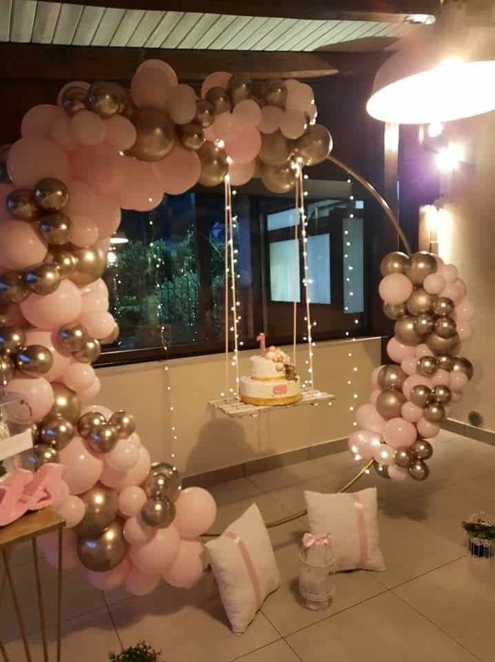 A large circular ring decorated with an arch of pink and metallic gold balloons for a birthday, anniversary, babyshower party. In the center, a small white swing adorned with fairy lights hangs, holding a birthday cake