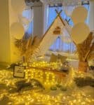 Romantic birthday canopy decor featuring a white teepee tent, fairy lights, white balloons, pampas grass, and an illuminated "HBD" sign on a white sheet with cushions.