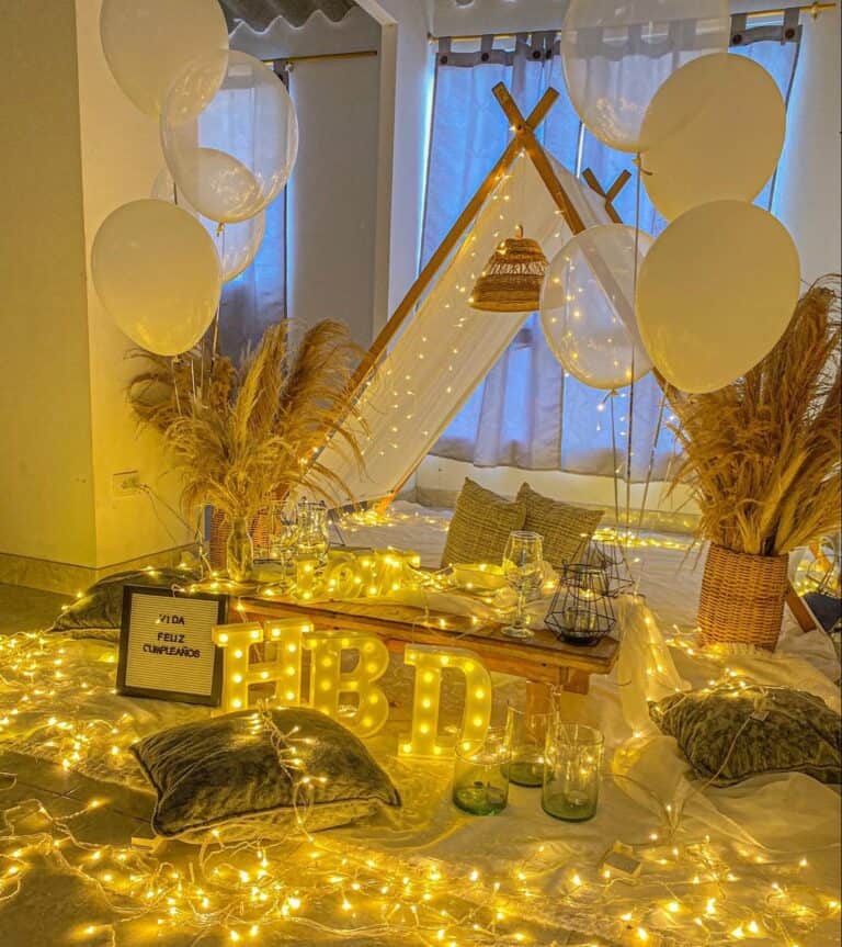 Romantic birthday canopy decor featuring a white teepee tent, fairy lights, white balloons, pampas grass, and an illuminated "HBD" sign on a white sheet with cushions.