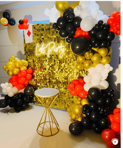 A festive party decoration featuring a gold sequin backdrop with a 'Let's Party' neon sign, a balloon garland in black, white, and red, and a geometric side table.