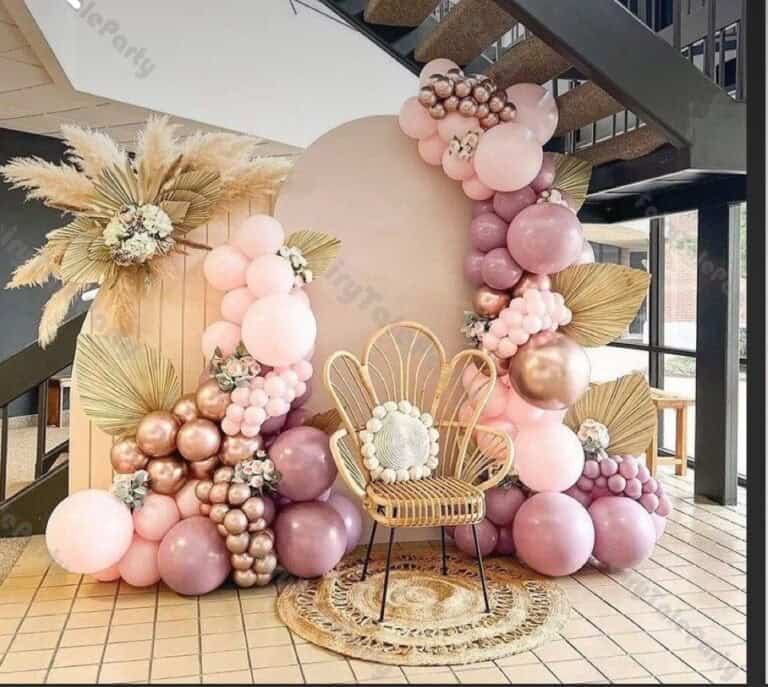 Pink and purple balloon party decoration featuring an organic balloon arch in shades of dusty pink, mauve, and metallic rose gold. The decor includes a stylish rattan flower-shaped chair, dried pampas grass, and large gold-painted palm leaves set against a circular beige backdrop.