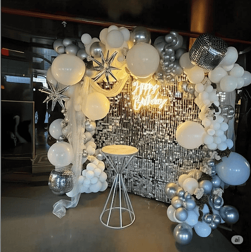 A stunning birthday party decoration featuring a shimmering silver sequin wall backdrop. A modern white neon sign that says "Happy Birthday" is centered on the wall. The backdrop is framed by an elegant arch of white and metallic silver balloons, with a large inflatable champagne bottle balloon on the right side.