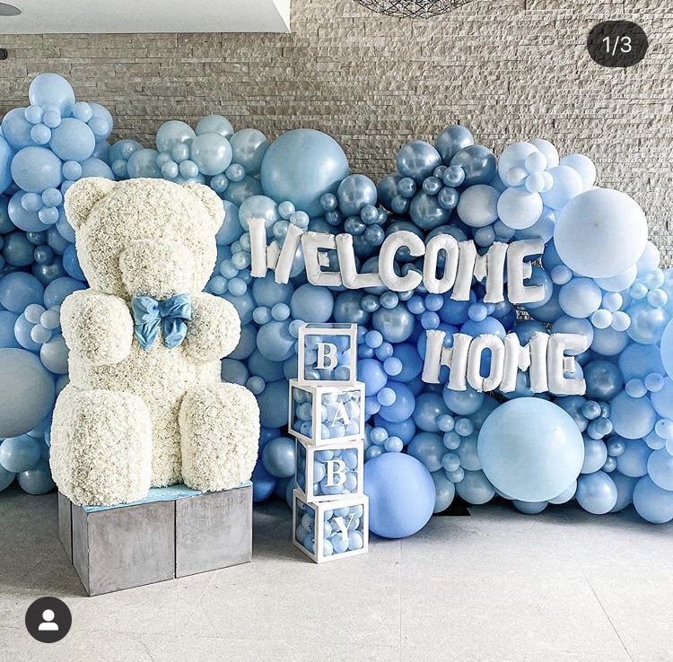 An elegant Welcome Baby Home decoration for a boy, featuring a large, white floral or moss teddy bear with a blue bow tie, placed in front of an expansive organic balloon wall. The wall is composed of various shades of light blue and white balloons, with white foil letters spelling out "WELCOME HOME" and stacked white boxes spelling "BABY."
