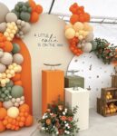 A Little Cutie Is On The Way baby shower decoration with an orange, sage green, and cream color palette, featuring organic balloon garlands, modern display pedestals, and floral arrangements.