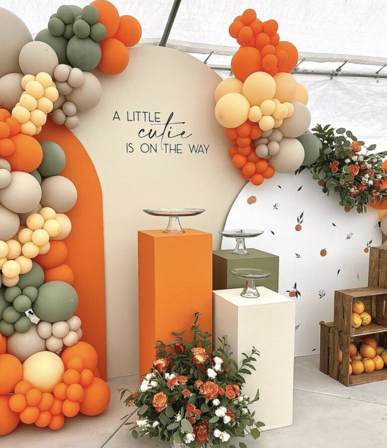 A Little Cutie Is On The Way baby shower decoration with an orange, sage green, and cream color palette, featuring organic balloon garlands, modern display pedestals, and floral arrangements.