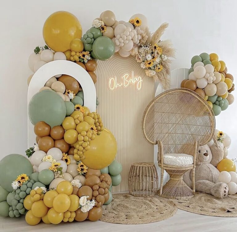 Boho-themed baby shower setup with a wicker chair, balloon garlands in earthy tones of mustard, sage, and beige, sunflowers, a teddy bear, and a neon sign reading "Oh Baby."