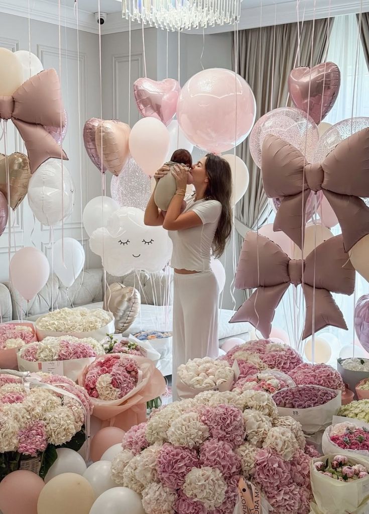 A joyful mother in white holds her newborn baby in a room lavishly decorated with a Baby Welcome Decorations Setup. The decor features large pink and white bow-shaped foil balloons, giant round pink balloons, heart-shaped balloons, and a cute cloud-shaped foil balloon. The floor is covered with dozens of stunning floral arrangements featuring pink and white hydrangeas and roses, creating an opulent Welcome Baby Home Decoration.