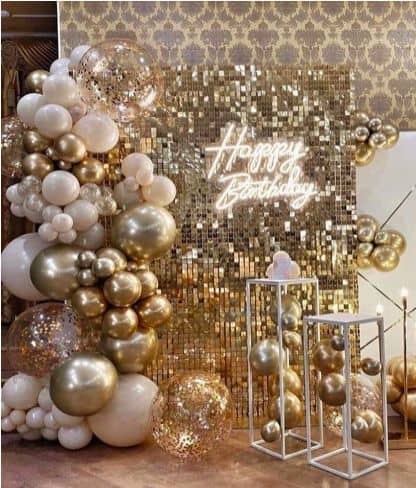 A luxurious birthday setup featuring a gold shimmer wall, a white neon "Happy Birthday" sign, and an elaborate balloon garland in white, beige, and chrome gold.
