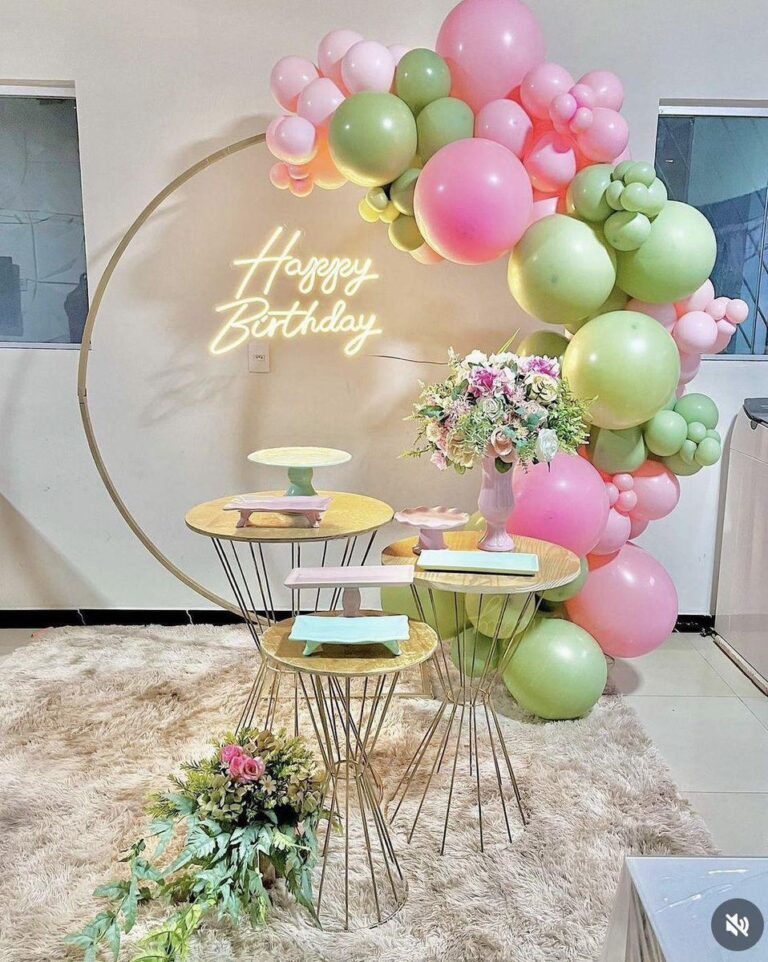 A modern birthday decoration setup featuring a circular gold metal frame with a "Happy Birthday" neon sign and an organic balloon garland in shades of pink and green. The setup includes multiple gold-toned wire dessert tables and a bouquet of flowers.