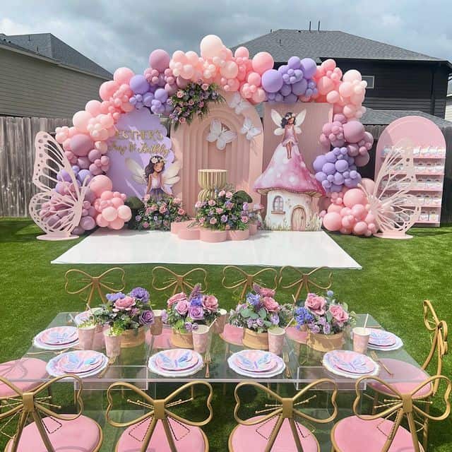 An enchanting outdoor fairy-themed first birthday party setup on a green lawn. The backdrop features a large pastel pink and purple balloon arch, cutouts of fairies, and large butterfly wings. In the foreground, a beautifully set dining table has pink chairs with gold bows and butterfly-themed plates.