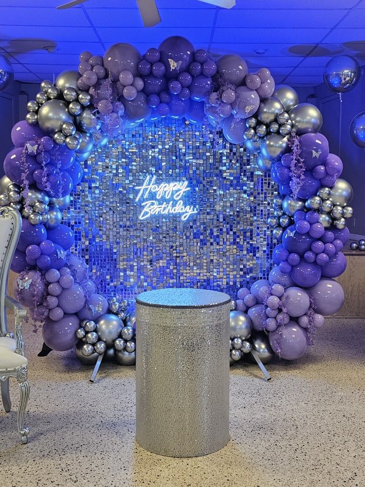 A large, circular balloon arch decoration for a birthday party. The arch is made of various sizes of purple, lilac, and silver chrome balloons, with some butterfly accents. In the center is a shimmering silver and blue sequin wall with a neon sign that says "Happy Birthday." In front of the wall is a cylindrical, silver sequin-covered pedestal. The overall color scheme is silver and shades of purple and blue, creating a festive and glamorous look.