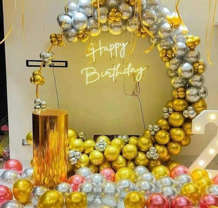 A luxurious circular balloon arch in metallic gold and silver for a birthday party, featuring a neon "Happy Birthday" sign in the center. The foreground includes a glossy gold pedestal and a large number '2' light-up marquee letter.