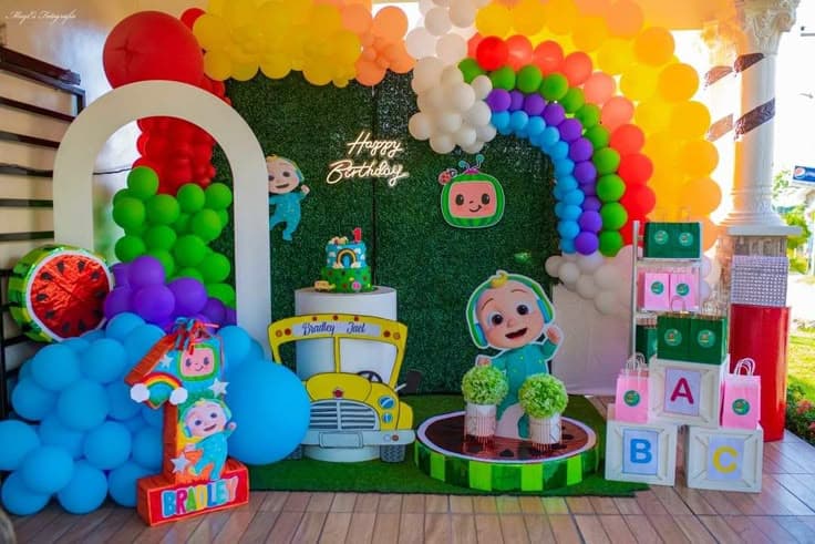 vibrant Cocomelon-themed birthday party decoration with a green grass-like backdrop, featuring a neon "Happy Birthday" sign, Cocomelon character cutouts, a rainbow balloon arch, a two-tier cake, and a number '1' prop with the name