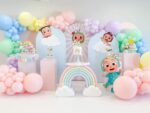 Cocomelon theme birthday decor featuring a pastel balloon arch, character cutouts of JJ, YoYo, and TomTom, and a rainbow cake stand. The setup includes beautiful balloon decoration in Gurgaon.