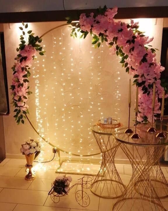 Elegant gold circular arch decorated with pink flowers and fairy lights, flanked by gold tables, creating a romantic anniversary room decor and backdrop.