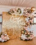 A luxurious anniversary decoration setup featuring a Glittering gold sequin wall as the backdrop. A glowing white neon sign on the wall reads "Crazy in Love." Framing the wall is an organic balloon garland in shades of gold, champagne, blush pink, and white, adorned with delicate pink flowers and greenery.