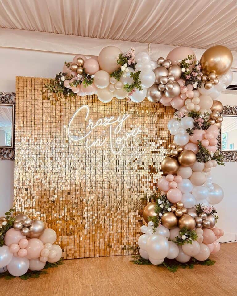 A luxurious anniversary decoration setup featuring a Glittering gold sequin wall as the backdrop. A glowing white neon sign on the wall reads "Crazy in Love." Framing the wall is an organic balloon garland in shades of gold, champagne, blush pink, and white, adorned with delicate pink flowers and greenery.