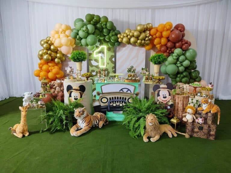 A lavish indoor kids' birthday decor setup with a safari or jungle theme, featuring a "Mickey Safari" twist. The backdrop includes white and green camo patterns, adorned with organic balloon decorations in shades of green, orange, and gold. In the foreground, there's a jeep facade, plush jungle animals like a lion, tiger, and giraffe, and dessert tables with Mickey Mouse safari elements. This elaborate custom theme decoration is perfect for a first birthday party setup.