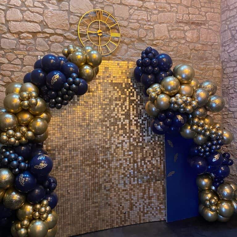 A luxurious anniversary decoration setup featuring a shimmering golden mosaic tile wall as a backdrop. Two elegant balloon garlands in a rich color scheme of navy blue and metallic gold frame the backdrop, forming a grand anniversary balloon arch. Above, a decorative gold clock adorns the rustic stone wall.
