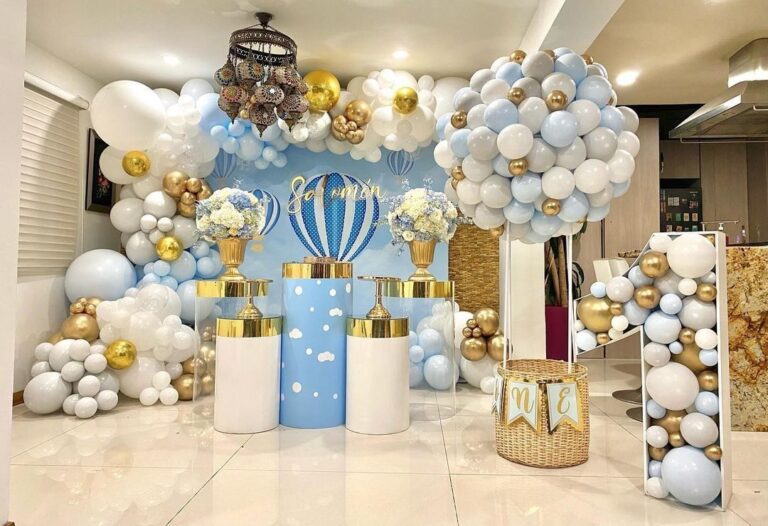 An elaborate first birthday decoration with a hot air balloon theme, featuring blue, white, and gold balloons, a large "ONE" sign, and clouds.