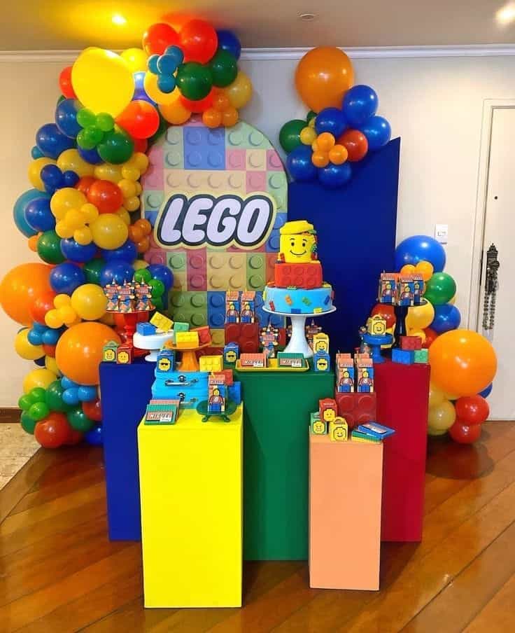 A vibrant LEGO-themed birthday dessert table with a large circular backdrop featuring the LEGO logo and colorful blocks. The table is adorned with a multi-tiered cake topped with a LEGO head, smaller cakes, numerous LEGO-themed treats, and various LEGO toy elements. Bright balloon garlands in red, blue, green, and yellow frame the display, which is set on colorful block-like pedestals.