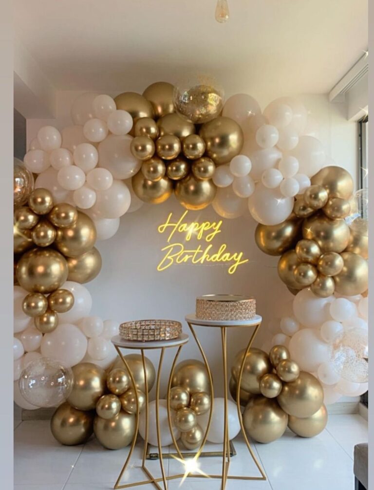 A stunning birthday party backdrop with a golden and white balloon arch surrounding a neon "Happy Birthday" sign, with two elegant gold dessert stands in front.