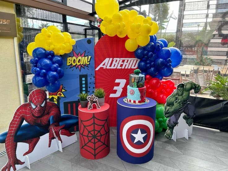 A superhero-themed 3rd birthday party setup for "Alberiu" featuring large cutouts of Spider-Man and the Hulk, a blue and yellow balloon arch, and themed pedestals holding a two-tier superhero cake and a small plant.