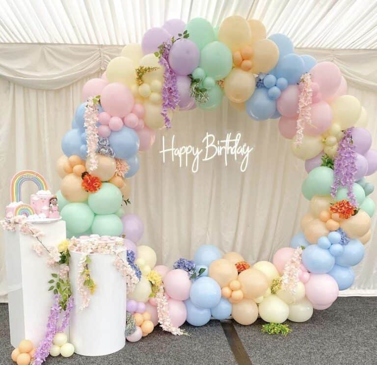 A large, circular balloon arch in pastel shades of pink, blue, yellow, green, and lavender, adorned with flowers. In the center, a white neon sign reads "Happy Birthday" against a white draped background. To the left, two white pedestals hold a small cake and other decorations.
