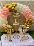 A luxurious, circular balloon arch in pink, white, and metallic gold framing a beige brick wall backdrop, featuring a neon 'Happy Birthday' sign, and three elegant gold and white display tables on a white faux fur rug.