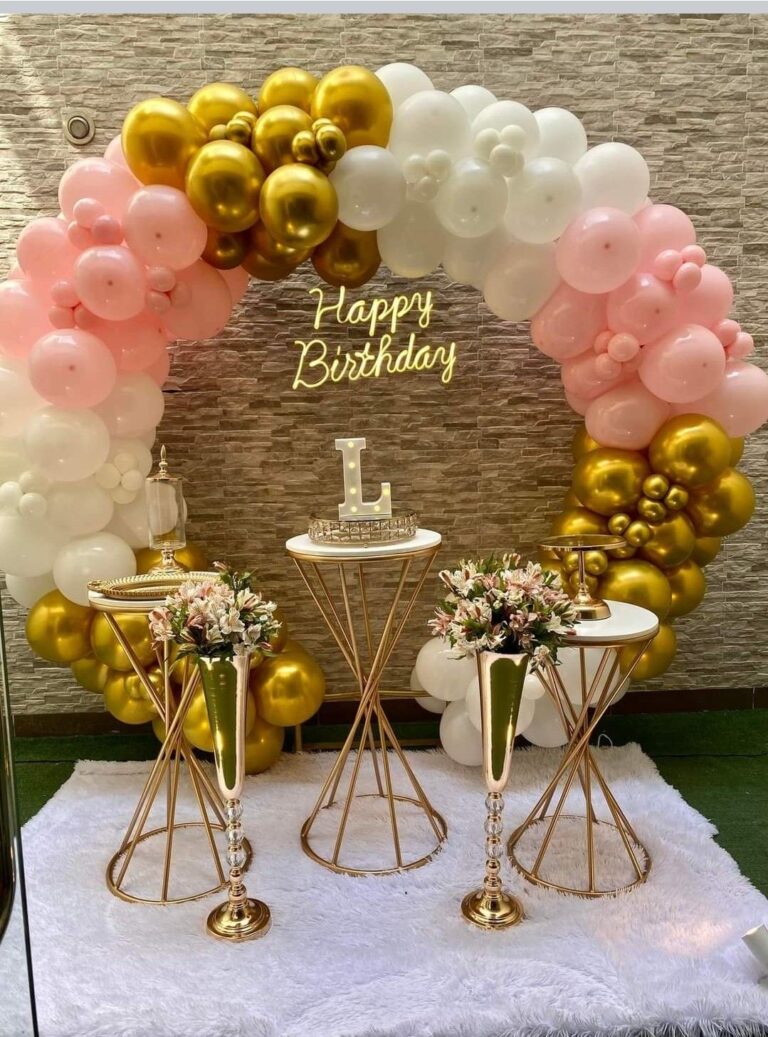 A luxurious, circular balloon arch in pink, white, and metallic gold framing a beige brick wall backdrop, featuring a neon 'Happy Birthday' sign, and three elegant gold and white display tables on a white faux fur rug.
