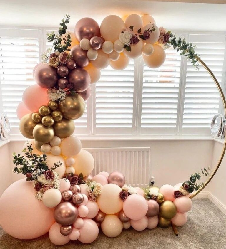 An elegant circular gold hoop balloon arch decoration with an organic arrangement of balloons in muted tones of dusty rose, mauve, chrome gold, and cream/peach. The arch is embellished with faux greenery, eucalyptus leaves, and small dark pink/burgundy floral accents, set up indoors in front of a window with white plantation shutters.
