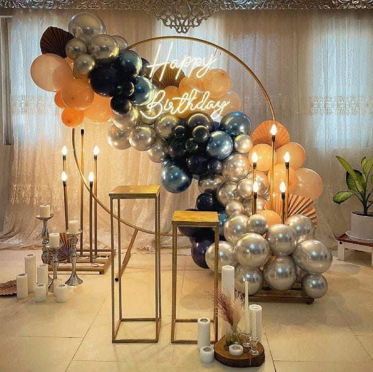 An elegant birthday party decoration featuring a large circular gold frame with a neon "Happy Birthday" sign. A cascading garland of silver, dark blue, and peach-colored balloons adorns the frame, flanked by tall golden candle holders and modern pedestal stands.