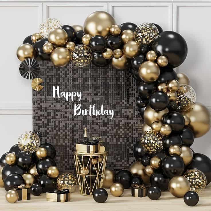 n elegant birthday decoration setup with a large black and gold balloon arch framing a dark sequin backdrop that reads "Happy Birthday." A gold wire table holds a champagne bottle and gifts, with more presents scattered on the floor.